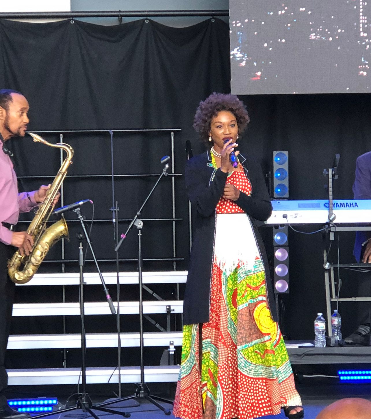 Buki singing on stage in a colorful African print dress, next to a saxophonist